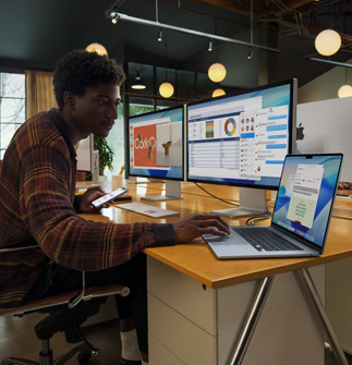 Una persona sentada en un escritorio trabaja con dos monitores Studio Display mientras usa una MacBook Air de 15 pulgadas, y los monitores muestran ventanas de Keynote, Vista Previa, Excel, Mensajes y Mail