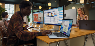 Una persona sentada en un escritorio trabaja con dos monitores Studio Display mientras usa una MacBook Air de 15 pulgadas, y los monitores muestran ventanas de Keynote, Vista Previa, Excel, Mensajes y Mail