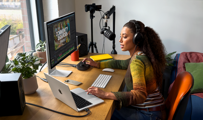 Una mujer en su casa trabajando desde su escritorio con una MacBook Pro conectada a dos monitores externos, usando audífonos y hablando en un micrófono